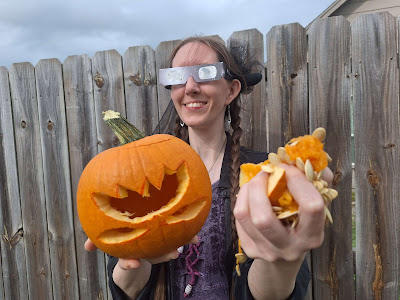 Gender fluid person in purple dress wearing solar viewing glasses presenting to the camera a finished jack-o-lantern and a fist full of pumpkin guts.