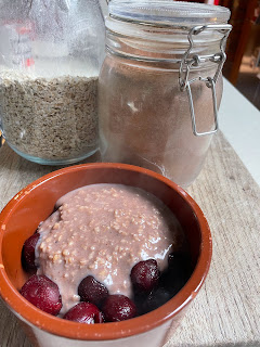 In the foreground is a ceramic bowl. Inside are some cherries, with pale brown porridge covering most of them. In the background are two Kilner jars. One has oats, the other is full of light brown powder.