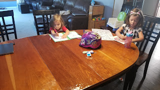 A photograph of two young girls at a large oval dining room table. They are coloring on some papers on the table.