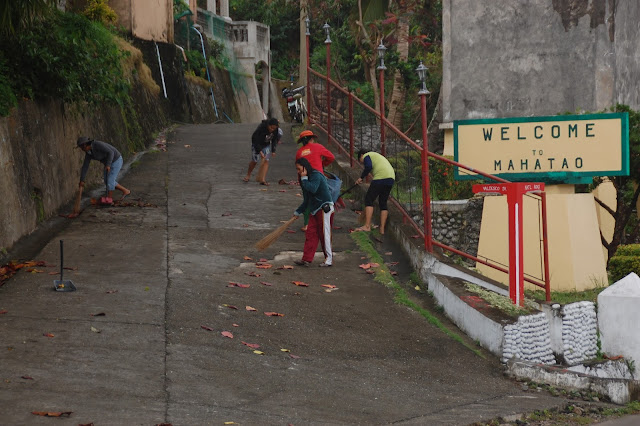 Five women sweep fallen leaves along an ascending street beside a Welcome marker in Mahatao, Batanes