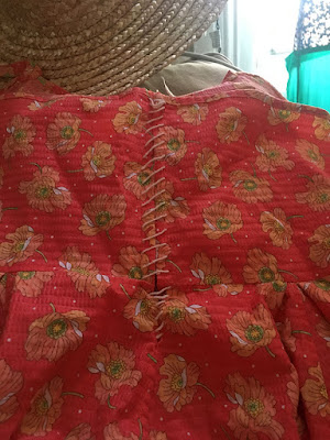 A close photo of the back of a coral sundress with large, scattered orange poppy motifs. White, slightly fuzzy cord laces the back edges together; at the top of the photo is the brim of a straw hat.