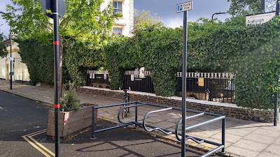 A view of the cycle parking which is a pale blue metal frame with horizontal parking hoops. There is a post sticking up with a "hire cycles" traffic sign on it.