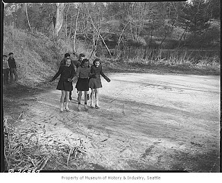 old black and white picture of five girls in skates on a frozen pond smiling for the camera