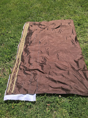 A big rectangle of dark brown silk laid out on grass in the sun, with a narrow band of gold embroidery running down the left edge and a piece of white fabric peeping out under that edge.