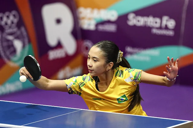 Giulia rebate a bola com o lado preto da sua raquete. Ela veste uma camiseta amarela do time brasil