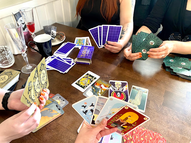 Pöydällä tarotkortteja. Neljä kättä pitelevät kortteja pöydän ympärillä.