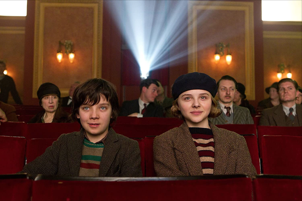Asa Butterfield and Chloe Grace Moretz seated in movie theater, grinning, directly facing out at the viewer