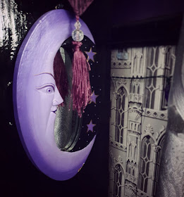 An oval mirror is on the right flank of a black-gloss book-case. The mirror frame depicts a lavender-coloured crescent 'man-in-the-moon' design with a black section with three stars painted on it. It matches the circular mirrors depicted in the picture above. There is a berry-purple tassel hanging partly over it from the altar cloth above, adorned with two clear crystal-effect beads. To the right of the image, the wall behind the book-case is visible, and a little of some greyscale wall-paper depciting Gothic architecture, it is a reproduction of an 1830's design and is in a block-print style.