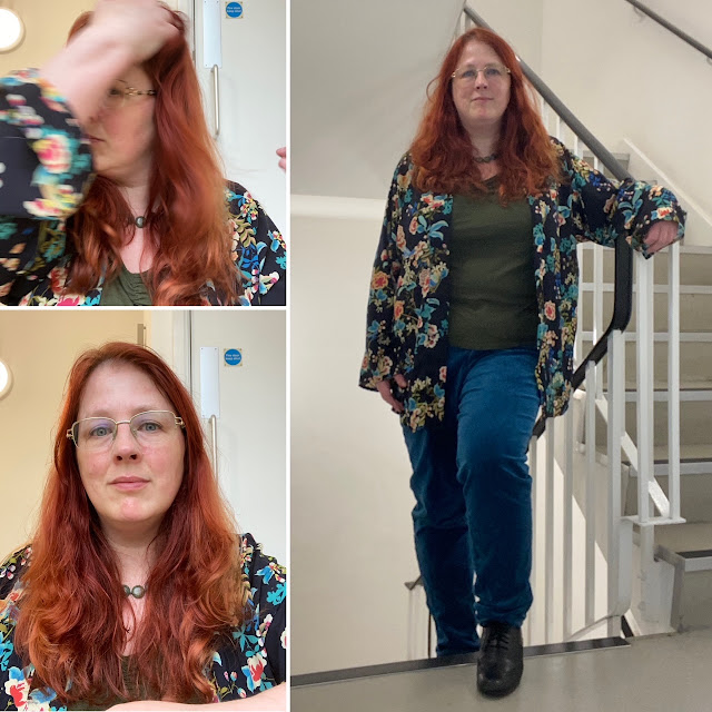 A redheaded white woman in teal jeans, an olive top, and a navy kimono top over it with a pattern of white, turquoise and salmon pink flowers and chaffinches.