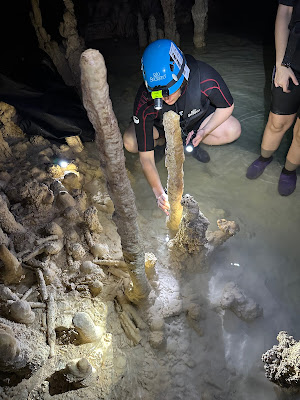 Woman in wetsuit with blue helmet and headlamp squatting in very shallow, clear water next to a skinny stalagmite in a cave