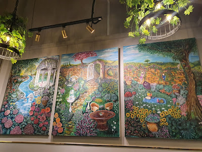 A triptych painted by Kadola featuring a garden, cats, and people with masks.