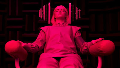 Young woman in institutional clothing with eyes closed, in a chair with futuristic machinery on both sides of her head