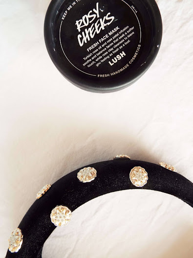 Close up picture of the Lush Rosy Cheeks Fresh Face Mask and black velvet pearled headband, in flatlay, on white bedding.