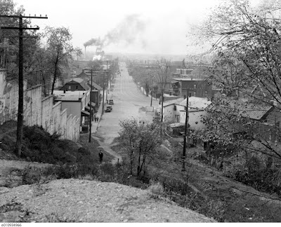 Black and white photo looking from the top of Nanny Goat Hill at the west end of Laurier (where the Slater ramp up to Bronson is now) down the hill and west along Albert and Wellington Street. At the foot of the viewer is a hill with various shrubs and a man walking up a narrow footpath through it. To the left is a concrete retaining wall with stepped panels following the hill. At the bottom of the hill is a stub of a street with buildings and two parked cars and a sidewalk on the left (south) side, beyond which Albert Street, bearing two sets of streetcar tracks, swoops in from the right and goes off straight away from the viewer. On either side of Albert Street is a sidewalk separated from the roadway by a boulevard, the one on the left planted with utility poles and the one on the right planted with trees. In the distance at the end of Albert Street is the silhouette of the railroad roundhouse, and to the left of it are smokestacks from various industrial facilities (including at least one brewery). I'm told this photo is from 1938 based on the presence and absence of various buildings in the background but the cars are all of an older vintage than other photos known to be from that year.