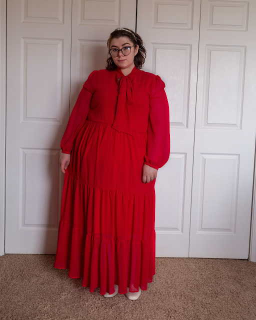 An outfit consisting of a red tie neck maxi dress with sheer bishop sleeves and white midi boots.