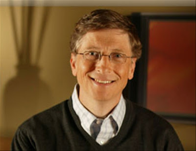 Descrição da foto: Bill Gates, homem de meia-idade, cabelos loiros escuros, curtos, pele e olhos claros. Na foro usa óculos de grau e está sorrindo tranquilamente olhando fixo para câmera. Veste uma camisa xadres e sobre ela um suéter escuro de lã; O fundo é em tons marrons com a sombra na parede de um vaso de flores do lado esquerdo.