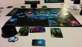A game of Abyss in progress. The board is painted to resemble an arena, filled with aquatic creatures looking into the central area, as seen from the last row of one the sections of the audience. Across the top are six spaces for cards; the leftmost is occupied by a deck of cards, with single cards in the next three spaces. The five sections of the audience on the other side of the 'arena' each has a space for cards, labelled with an icon representing the five races of allies: jellyfish, crabs, seahorses, shellfish, and squids. Of these spaces, only the crabs and the seahorses do not have a stack of cards on them. Along the bottom of the board is a track of seven spaces for cards. The leftmost is occupied by a deck, and the three rightmost spaces each have a single face-up card illustrated with a lavish painting of a fish-man. Around the board are various items (cards, tokens, sea-shell-shaped cups to hold pearls, etc) belonging to the various players.