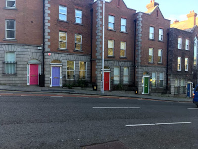 A series of narrow, three-storey buildings with brightly painted doors in the grey stone ground floor, with red brick above, stepped to follow the downward slope of the street. The doors are bright pink, clear lavender, fire engine red, pine green, and ocean blue.