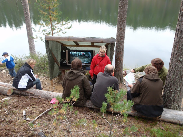 Neljä ihmistä istuu ja katsoo kahta muuta ja telkkaria metsässä, taustalla lampi