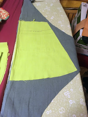 A large, nearly trapezoidal piece of bright lime-green fabric, taller than it is wide and with a gently curved lower edge, laid out on a shallow triangle of muted teal fabric where it just fits, all on a pale green floral ironing board.