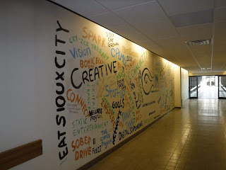 this mural in the Sioux City Skywalk system was painted as part of an event called Innovation Market and features inspiring words like "spark" and "goals" in black, orange, spring green, and sky blue on a white wall in the Sioux City Skywalk system