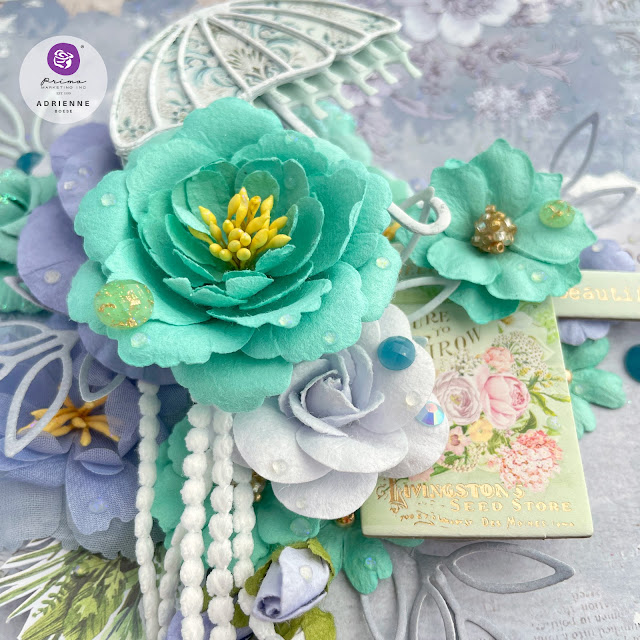 Mixed media panel on paper. Blue and green flowers and papers with a die cut umbrella and leaves, ephemera and DIY raindrop medium created with Finnabair soft gloss gel and glitter.