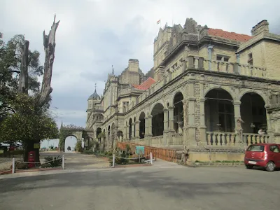 A grand colonial-era building with arched verandas, intricate stone architecture, and red-tiled roofs, known as the Viceregal Lodge in Shimla, Himachal Pradesh, India. A red car is parked nearby, and a tree with yellow flowers stands at the entrance.