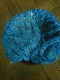 a blue lace shawl live on a circular needle, tucked into a wooden yarn bowl.  There is a stitch marker with a shell charm clipped into the work.