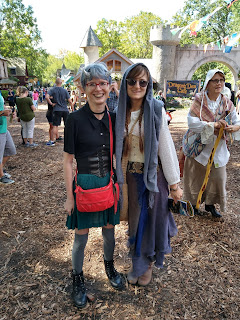 A girl dressed as a demon and a girl dressed as a peasant standing together. There is an old crone in the background. They are standing in a wooded area in front of a castle.