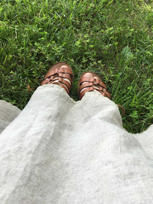 The hem of a light-grey linen dress, with brown-sandaled white feet peeping out on the lush grass.