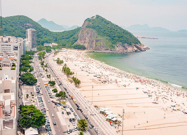 spiaggia affollata vista dall'alto, a sinistra la città, a destra il mare, al centro la strada con le auto, sullo sfondo bellissime montagne verdi