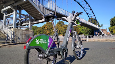 A green, grey and purple hire cycle sits in front of a bowstring style steel bridge crossing a large road.