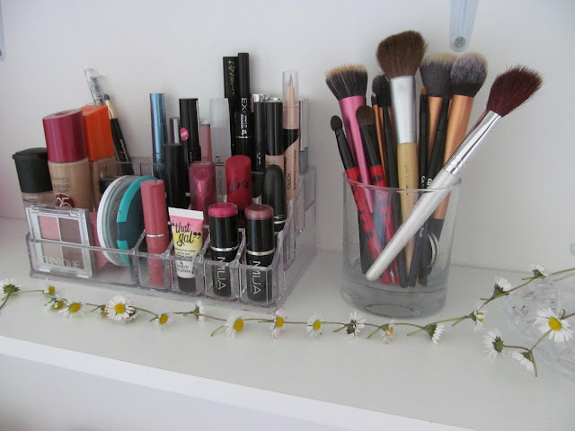 acrylic makeup stand surrounded by daisy chain with makeup brushes on right in clear candle jar