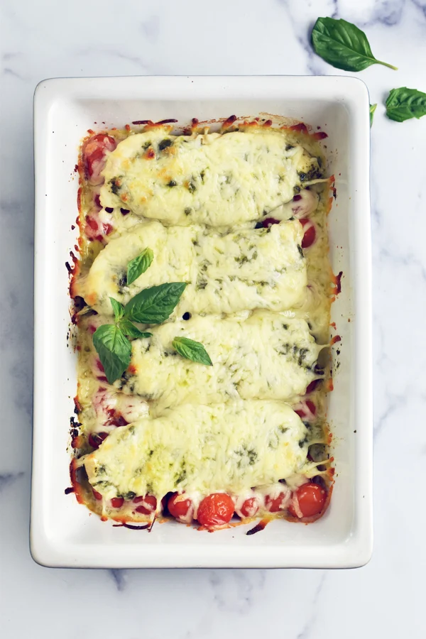 A tempting casserole dish filled with baked chicken pesto, generously covered in melted mozzarella cheese and beautifully garnished with fresh basil leaves, ready to be devoured and enjoyed.