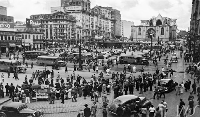 A fotografia, em preto e branco, mostra a Praça da Sé, no centro de São Paulo. Está lotada de pessoas e há alguns carros e ônibus. No fundo, há alguns prédios e a Catedral da Sé em construção.