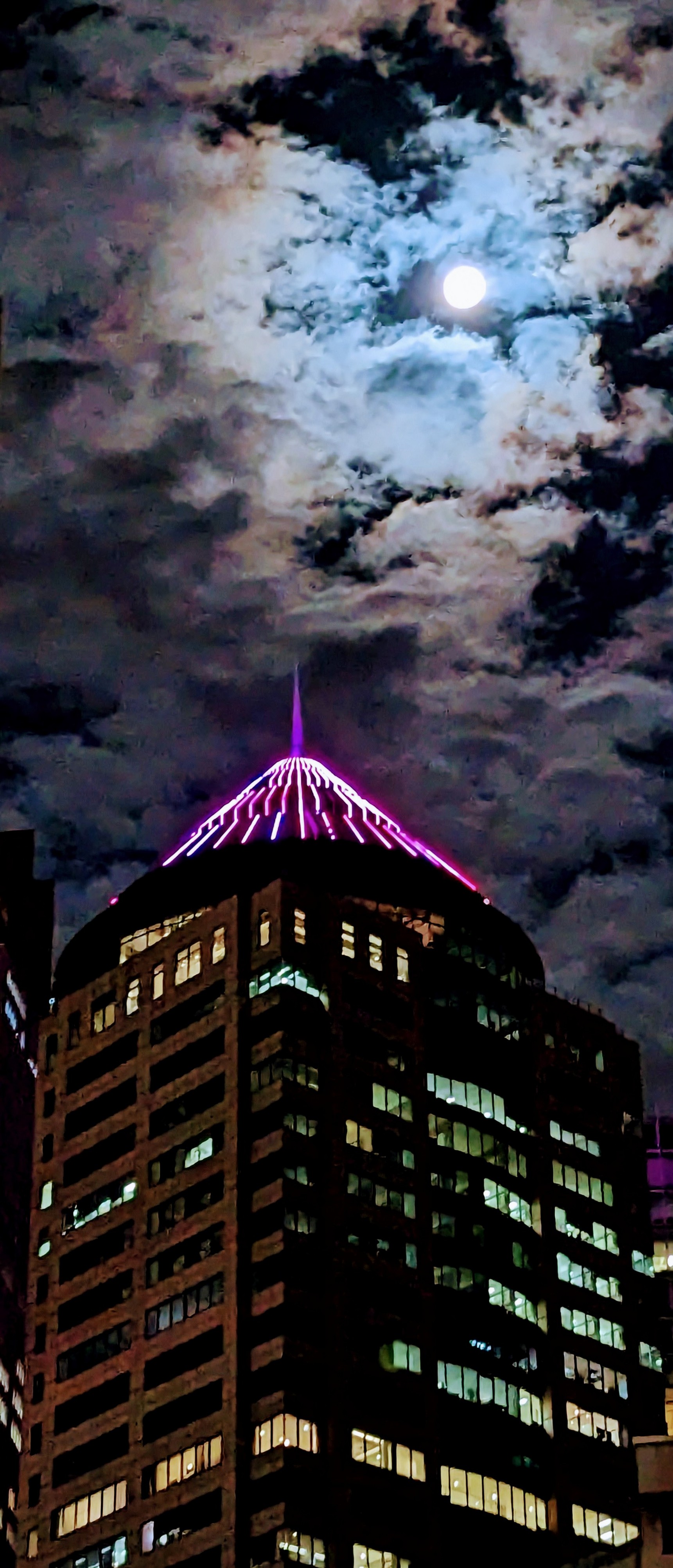 Sydney high rise with pink lit up conical top and arial underneath a bright full moon that lights up the surrounding clouds.