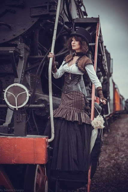 Woman dressed in steampunk clothing, on the side of a steam train. Striped trumpet skirt, corset, bolero jacket, top hat, parasol.