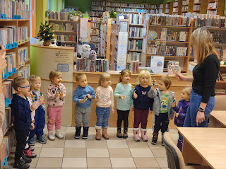 Tło: sala biblioteczna. Przed ladą biblioteczną stoją dzieci w ręku trzymają małe metalowe grzechotki. Obok dzieci stoi pani przedszkolaka. Na ladzie stoi choinka ze złotymi lampkami.