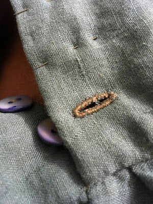 A close-up of a tan buttonhole worked densely in muted teal fabric at the base of a deep button placket, with pins marking the positions of more buttonholes and two blue-shell buttons showing on the underlap.