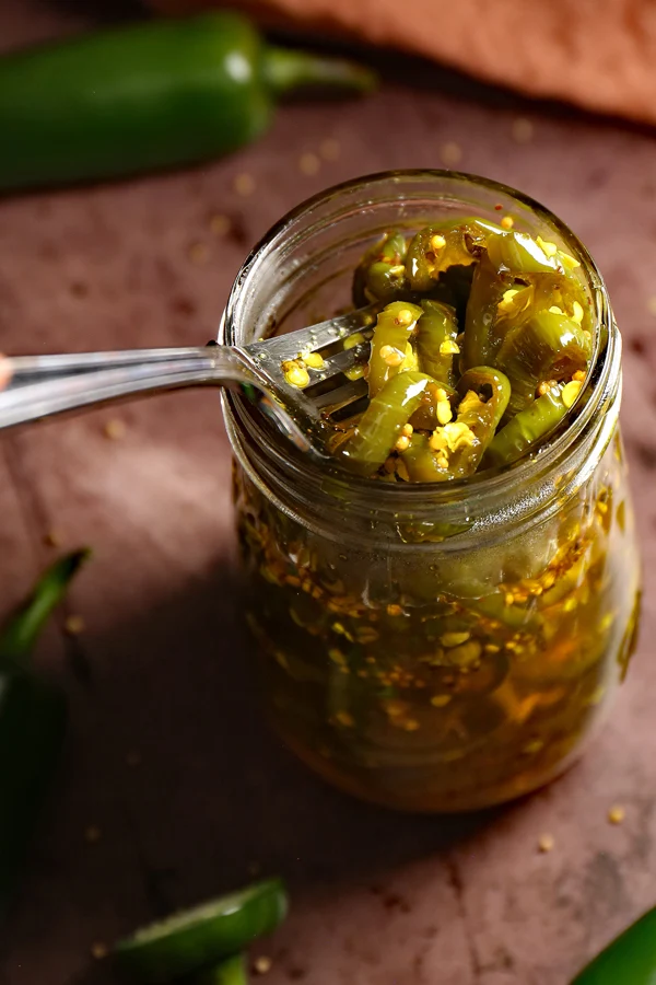 A close-up shot of a jar filled with delectable cowboy candy. A fork is reaching into the jar, skillfully picking up the tantalizing candied jalapenos. The vibrant colors and sticky textures of the jalapenos promise a delightful blend of spiciness and sweetness for a truly mouthwatering experience.