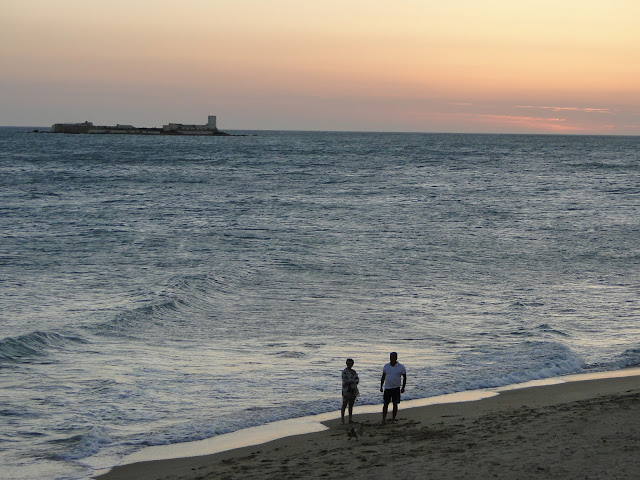 Playa con una pareja en la orilla con el mar con oleaje y una pequeña isla al fando.