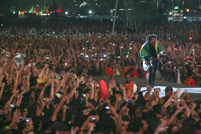 Foto do vocalista do Foo Fighters, Dave Grohl, na passarela em meio ao público em show no Brasil. Vários braços esticados no público, com câmeras e celulares fotografando tudo