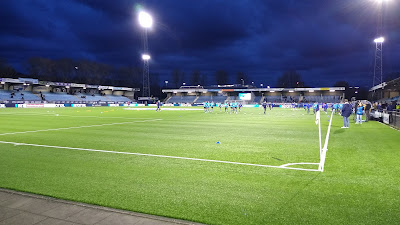 The view across a flood lit football pitch with stands around it.