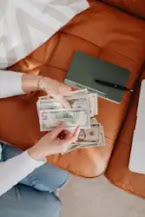 An entrepreneur sits on the floor counting money while resting her hand on the sofa with a notebook and a pen.