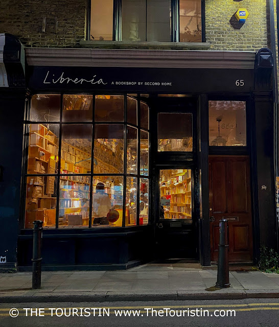 Through the illuminated bookshop window, piled bookshelves can be seen snaking along the walls on both sides of the elongated room all the way up to the ceiling. The words 'Libreria. A bookshop by second home' on its black facade in white letters.