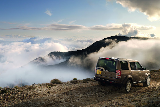 Rear 3/4 view of bronze 2011 Land Rover LR4 on dirt trail above mountains with fog and clouds