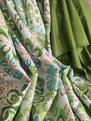 Two large, draped pieces of fabric, one with large Hawaiian-print style flowers and leaves in two shades of green, turquoise, and grey, and the other light lime-green with small, densely scatted gold dots.