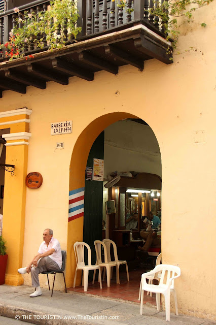 A grey haired man sitting on a chair in front of the yellow facade of a hairdresser's