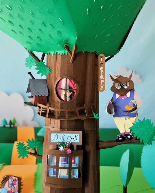 detailed diorama scene made of paper showing owl perched on tree limb, birdhouse and book nook shop inside tree trunk