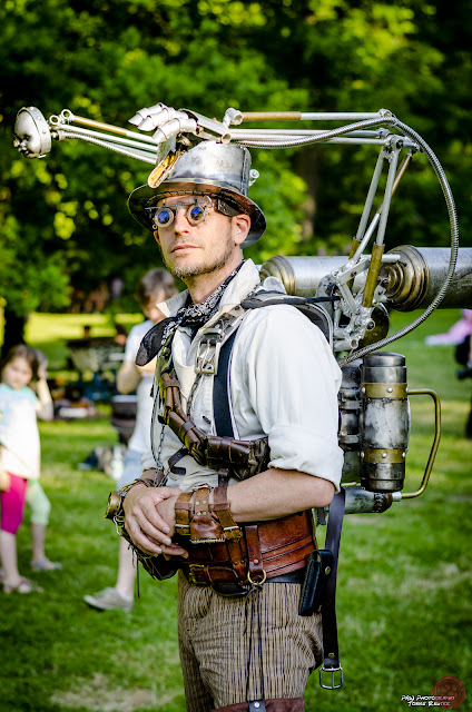 Steampunk man wearing a strange gadget/contraption strapped to his back. Mechanical arm and hand, plus baby doll head